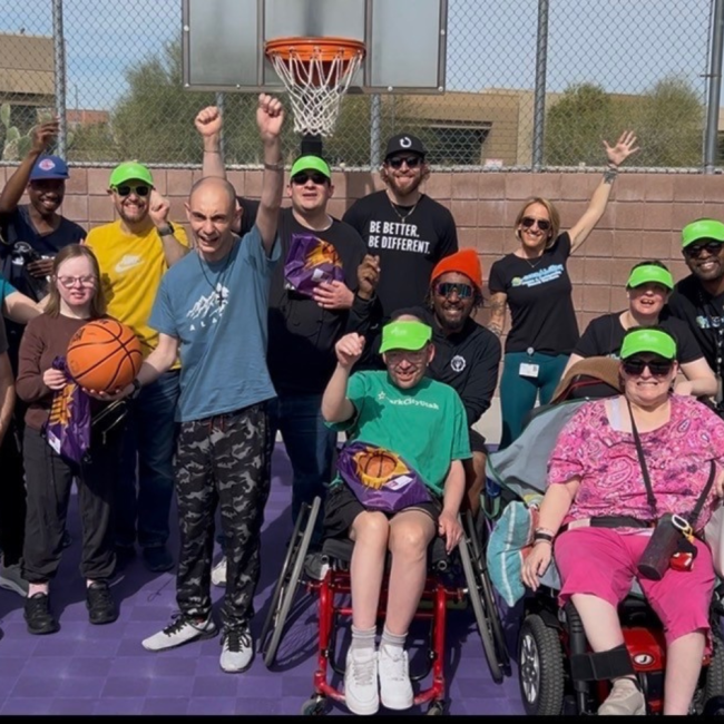 A diverse group of people, some in wheelchairs, pose and smile on an outdoor basketball court, with a hoop visible in the background. Several participants wear green caps. A diverse group of people, some in wheelchairs, pose and smile on an outdoor basketball court, with a hoop visible in the background. Several participants wear green caps.