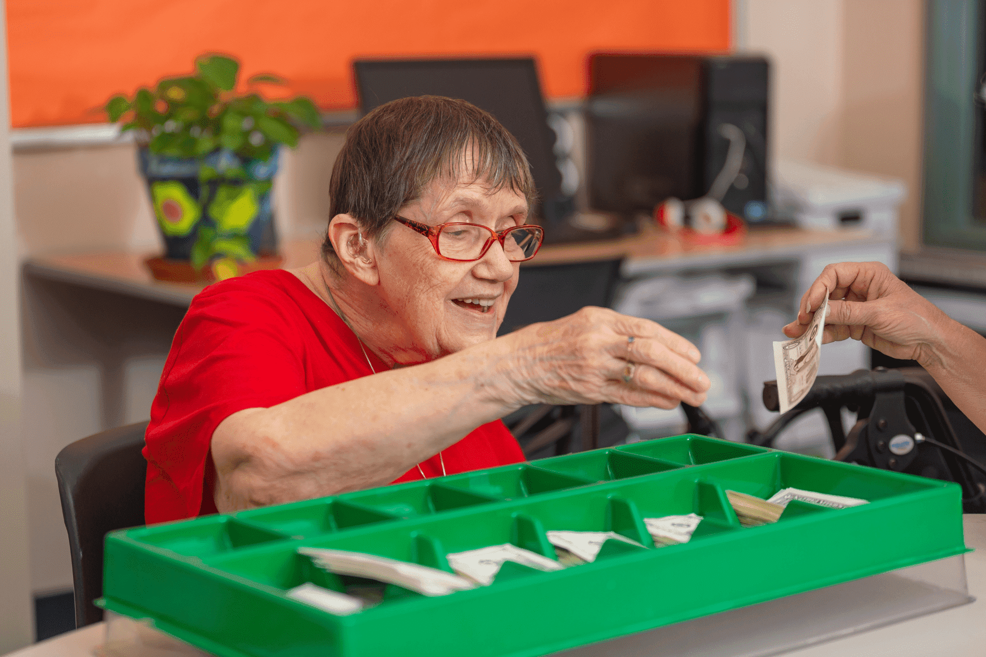 United Cerebral Palsy Home 9 An elderly woman with glasses hands money to someone across a green tray with divided sections, in an early learning center setting with computers in the background.