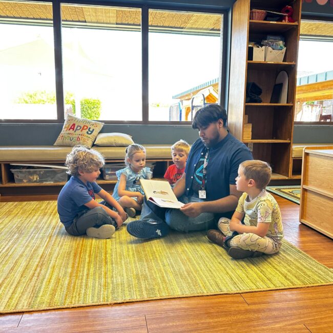 An adult reads a book to four young children sitting in a circle on a rug in a classroom with large windows. An adult reads a book to four young children sitting in a circle on a rug in a classroom with large windows.