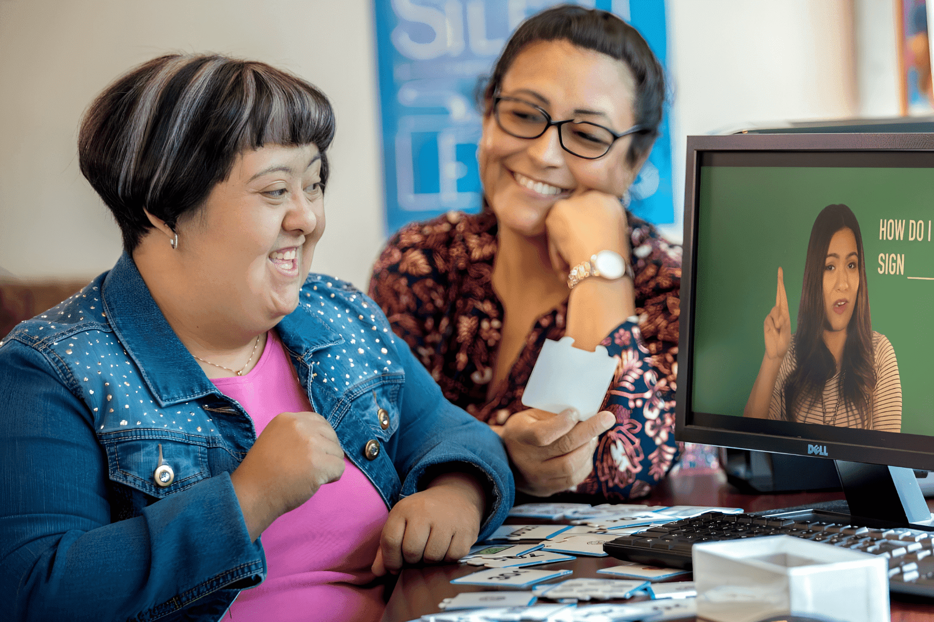 United Cerebral Palsy Home 8 Two women sit together at a computer, smiling, while watching a sign language lesson video and holding flashcards.