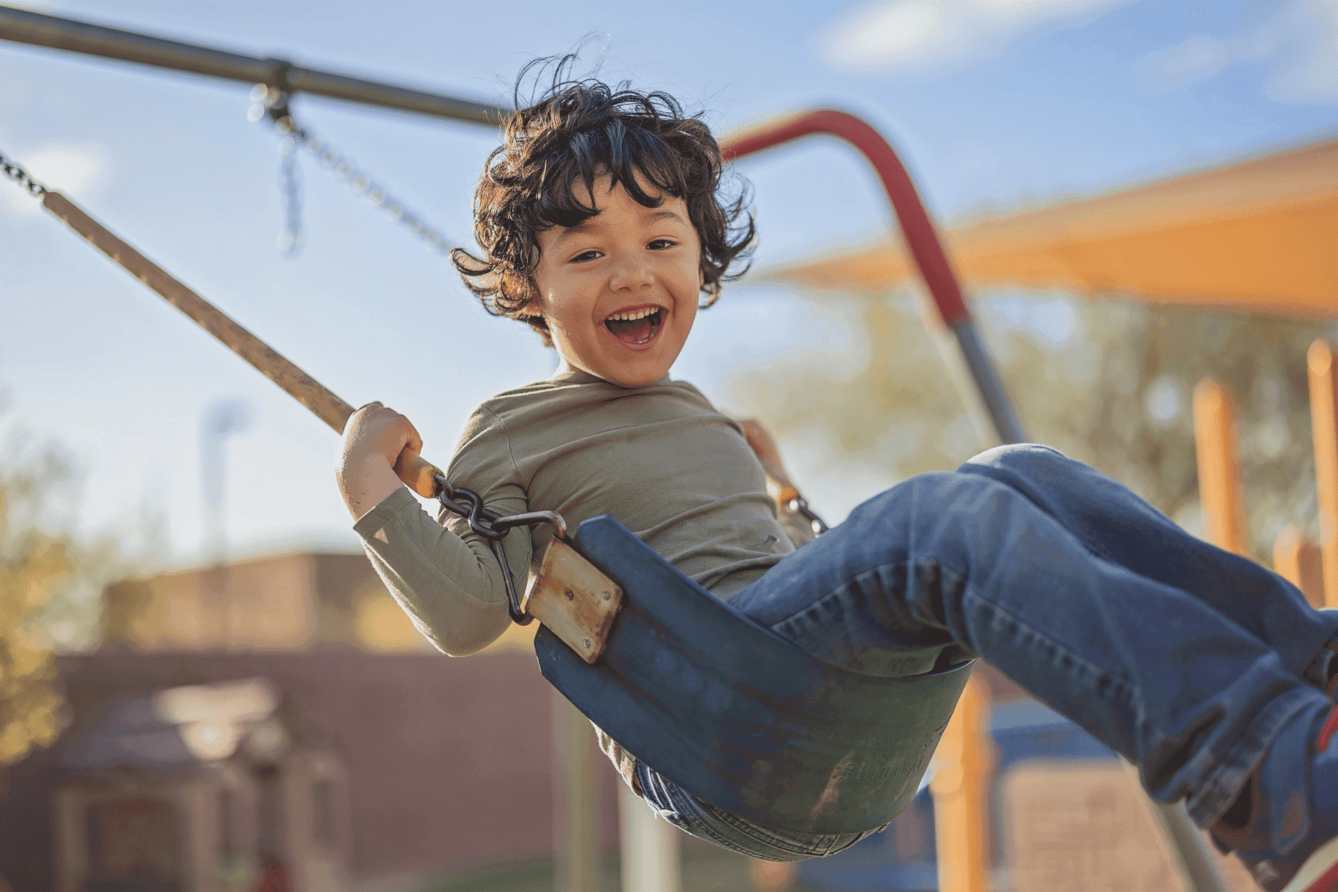 United Cerebral Palsy Home 6 A young child with curly hair smiles while swinging at a playground on a sunny day.