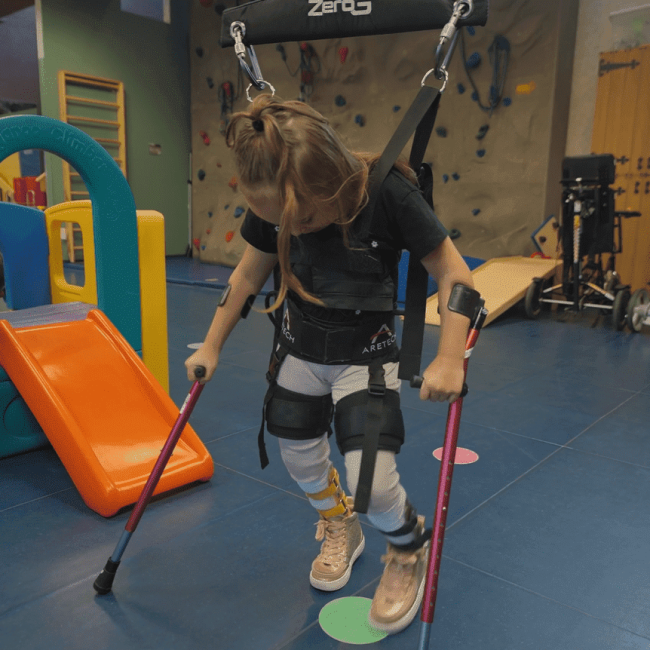 A young girl in a harness uses crutches for support in a therapy gym, with play equipment and a climbing wall in the background. A young girl in a harness uses crutches for support in a therapy gym, with play equipment and a climbing wall in the background.