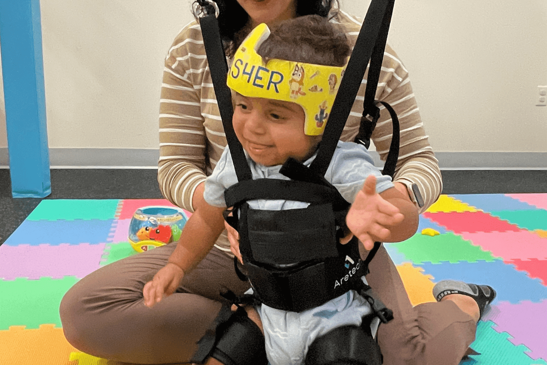 United Cerebral Palsy Home 7 A young child wearing a yellow headband and harness sits on a woman's lap on colorful foam mats in a playroom.